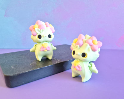 Two small figurines designed to look like pixies with flower crowns, one in mint color with a bee design on its chest, and the other in a similar style with a different pattern.