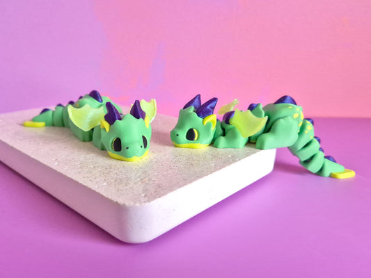 Small green dragon figurines with lime and metallic navy accents, with or without wings, positioned on a pink and purple background.