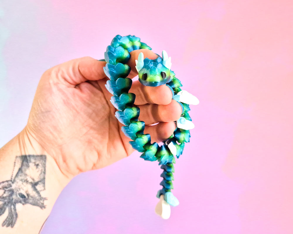 Hand holding a blue and green dragon-shaped fidget toy against a pink and purple gradient background.